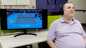 Blind man sitting in a chair wearing headphones, with a monitor behind him playing a tennis broadcast video.